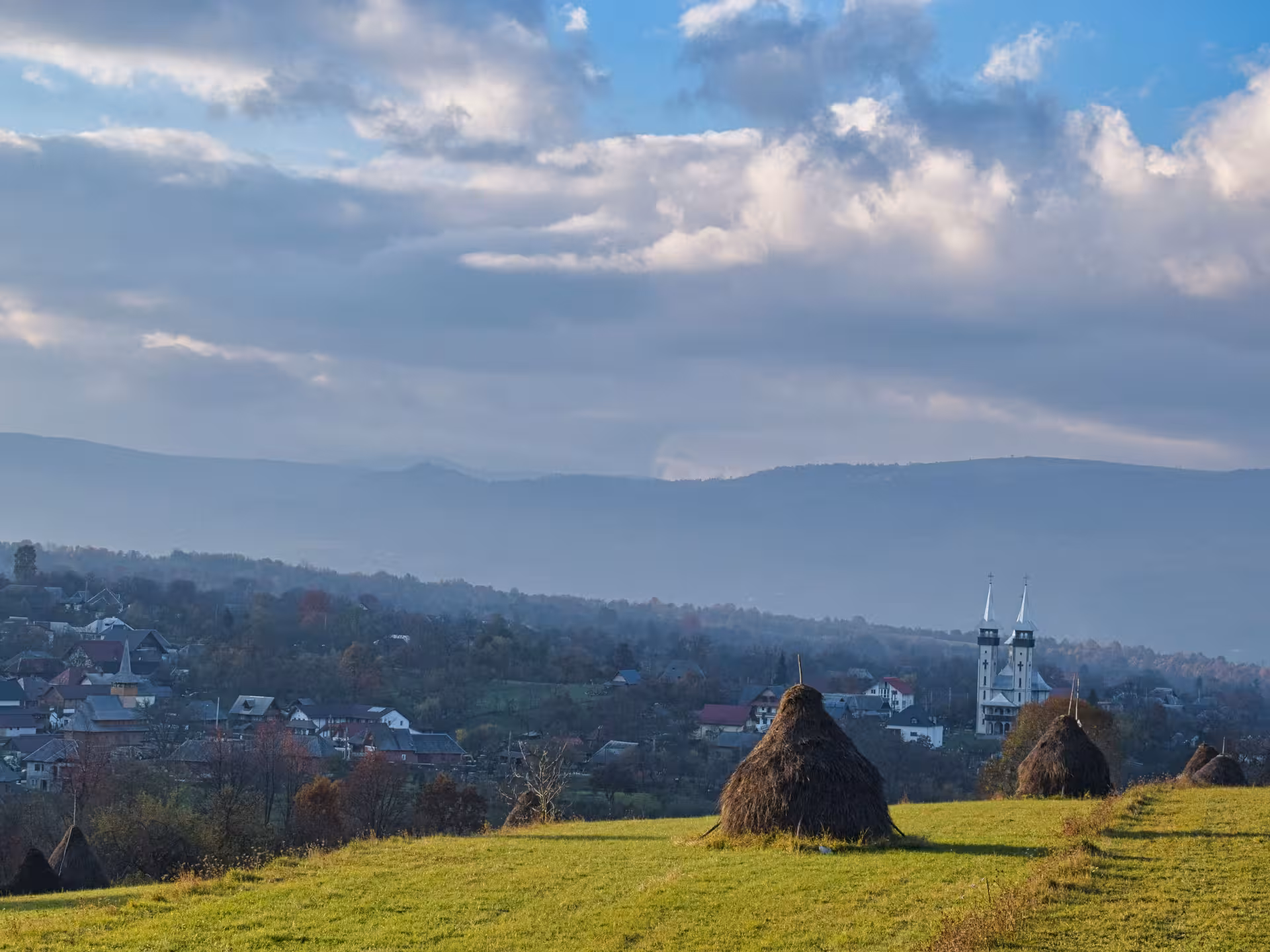 Haystack-dotted green meadow overlooking a Maramures village with twin-spired church and misty Carpathian mountains