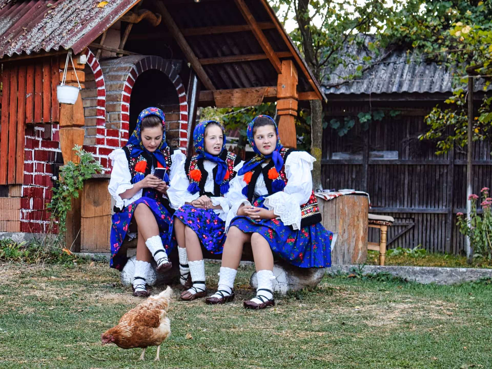 Three young women in colorful traditional Maramures folk costumes sitting together in an authentic Romanian village courtyard