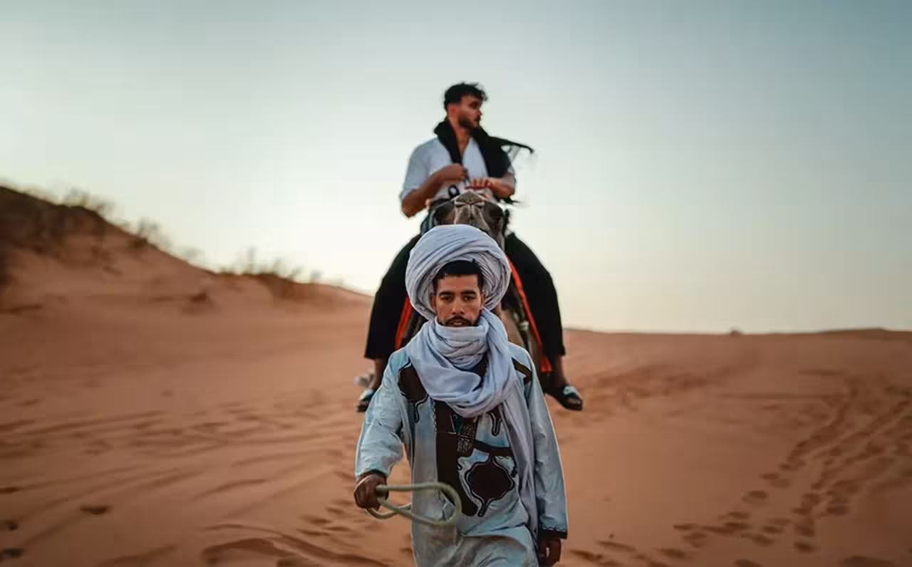 A guide leads a camel carrying a tourist through the captivating sands of Erg-Chebbi on a Marrakech to Fez tour.