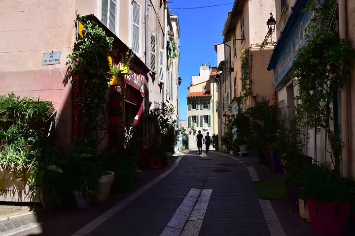 Narrow sunlit alley in Marseille Old Town with colorful buildings, lush plants, and two people walking