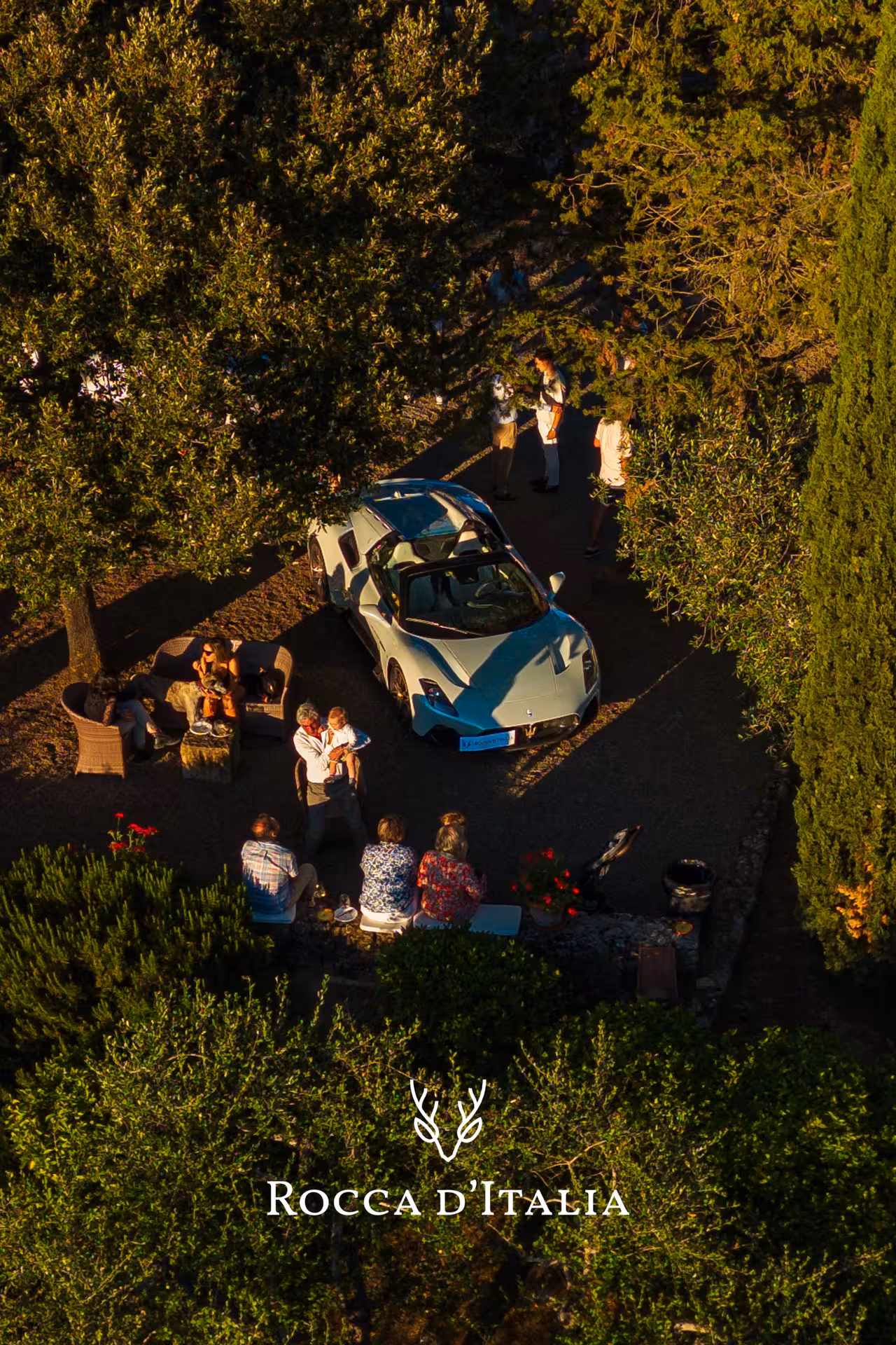 Aerial view of a light blue Maserati supercar surrounded by guests at a Tuscan garden wine tasting event