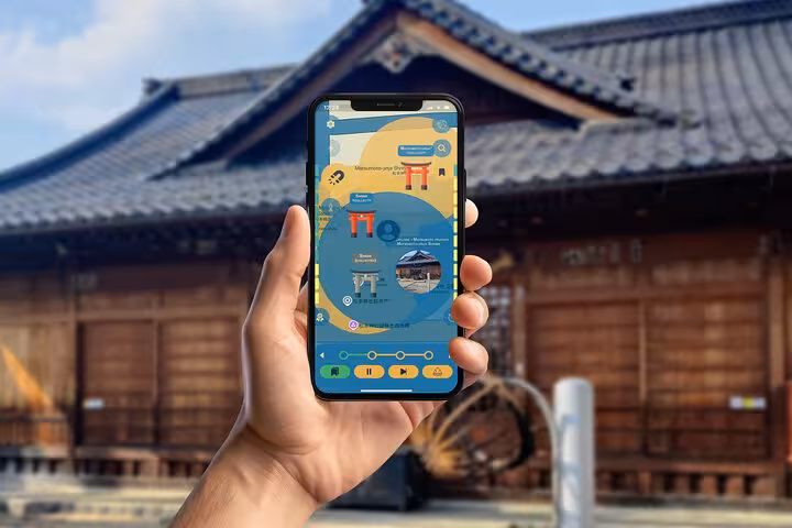 Hand holding smartphone with Matsumoto self-guided walking tour app open in front of a traditional Japanese wooden shrine building
