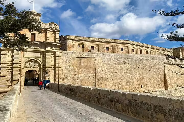 Impressive stone gateway entrance to Mdina, Malta, with tourists exploring the ancient city walls under a bright blue sky.