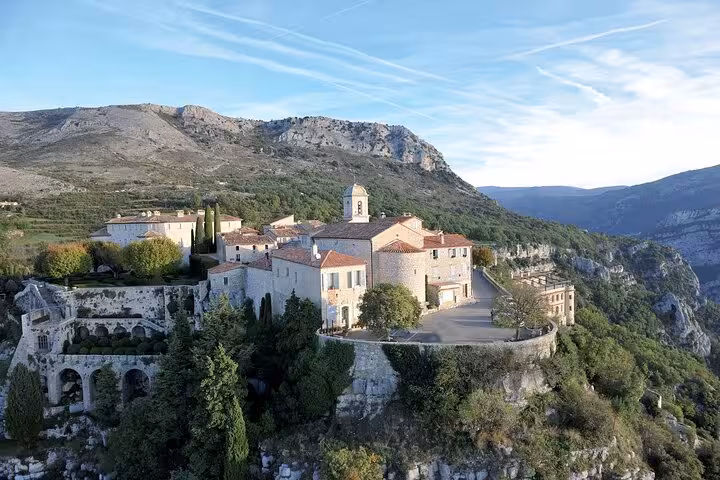 Aerial view of a medieval village perched on a cliff in the Provencal countryside, surrounded by lush hills.