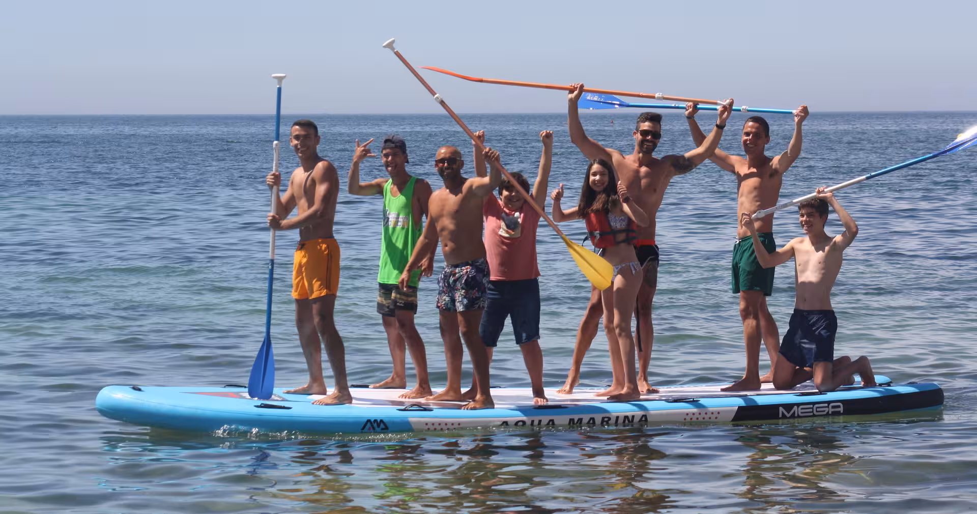 A cheerful group enjoying the Mega Paddle tour on an Aqua Marina giant SUP board in calm ocean waters.