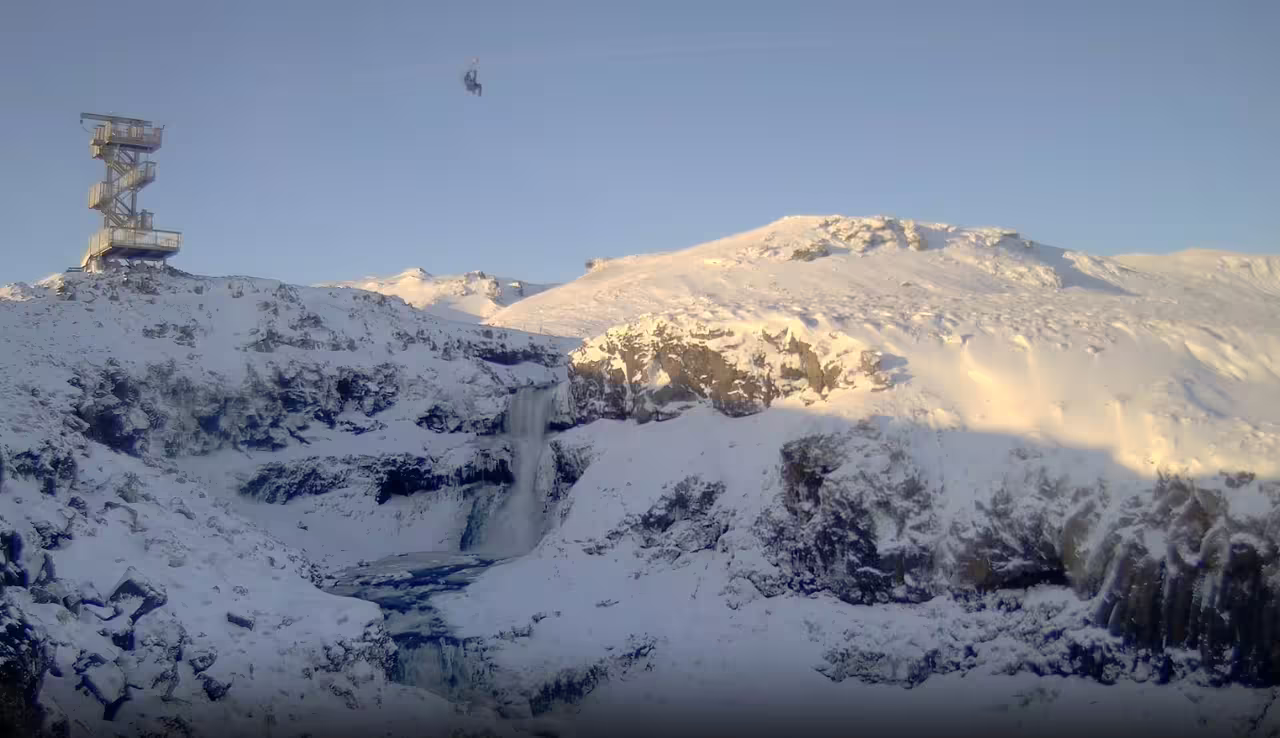 Adventurer on Mega Zipline ride above snowy Icelandic landscape and waterfall, with transfer from Reykjavik included.