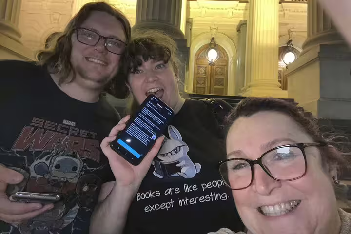 Three smiling friends on Melbourne night walking tour holding phone with puzzle clues outside historic building