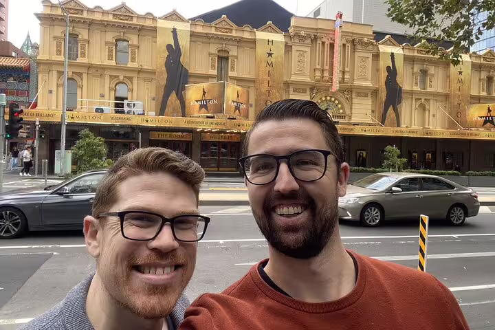 Two smiling men in glasses taking a selfie in front of the Hamilton musical theatre in Melbourne