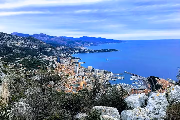 Aerial view of Monaco coastline with harbor, dense city buildings, and blue Mediterranean Sea backed by mountains