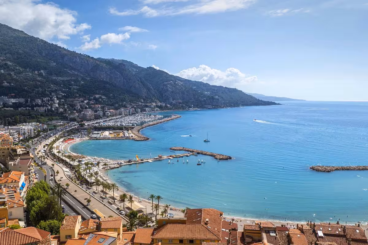 Aerial view of Menton's curved coastline with turquoise Mediterranean sea, palm-lined promenade, marina, and hillside town