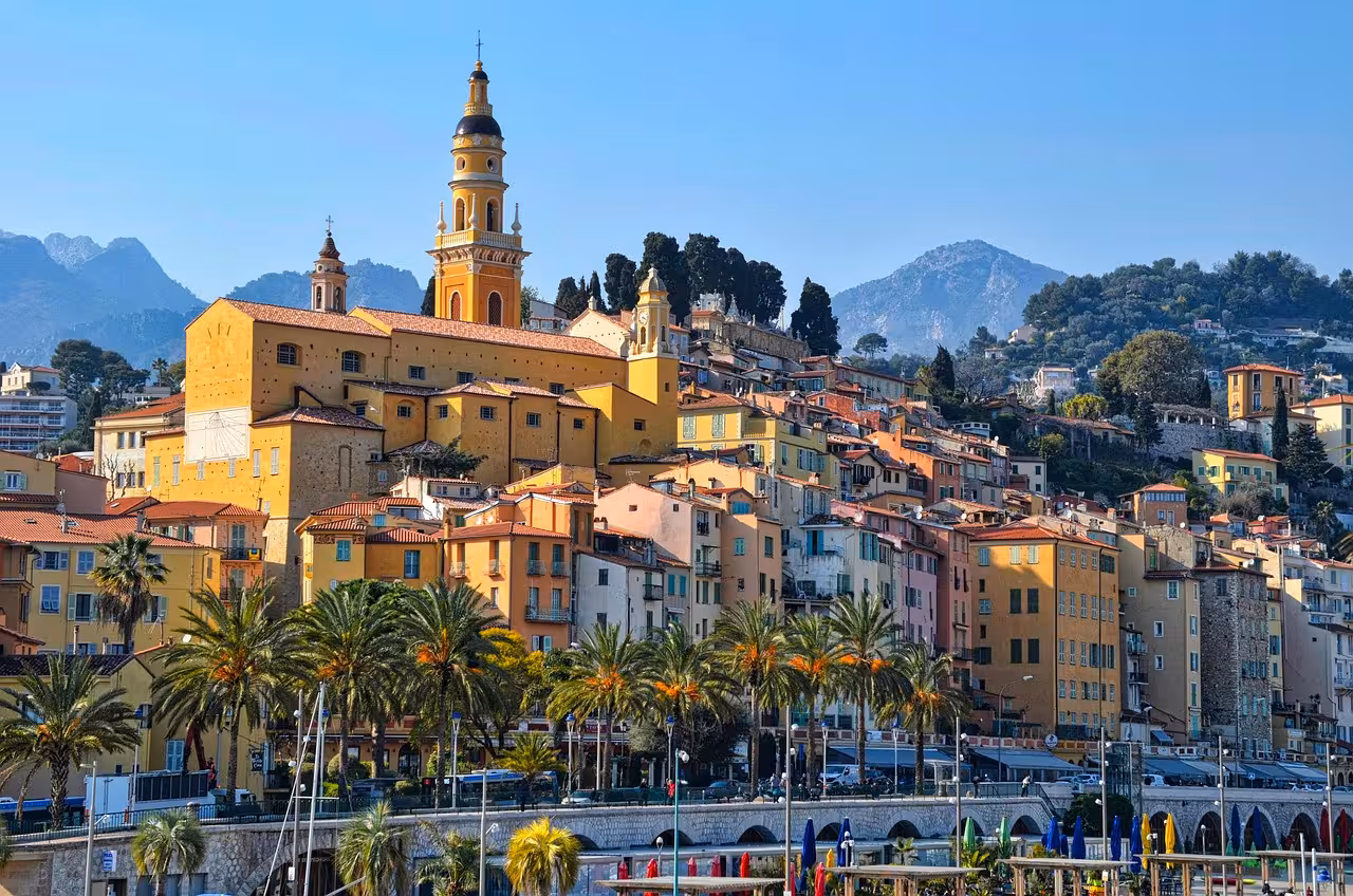 Colorful hilltop old town of Menton France with baroque church tower, palm trees, and Mediterranean mountains