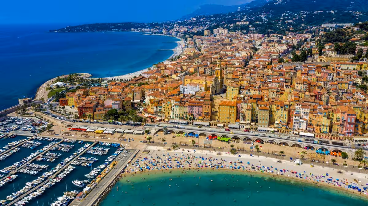 Aerial view of Menton's colorful old town, marina with white boats, and crowded beach on the French Riviera