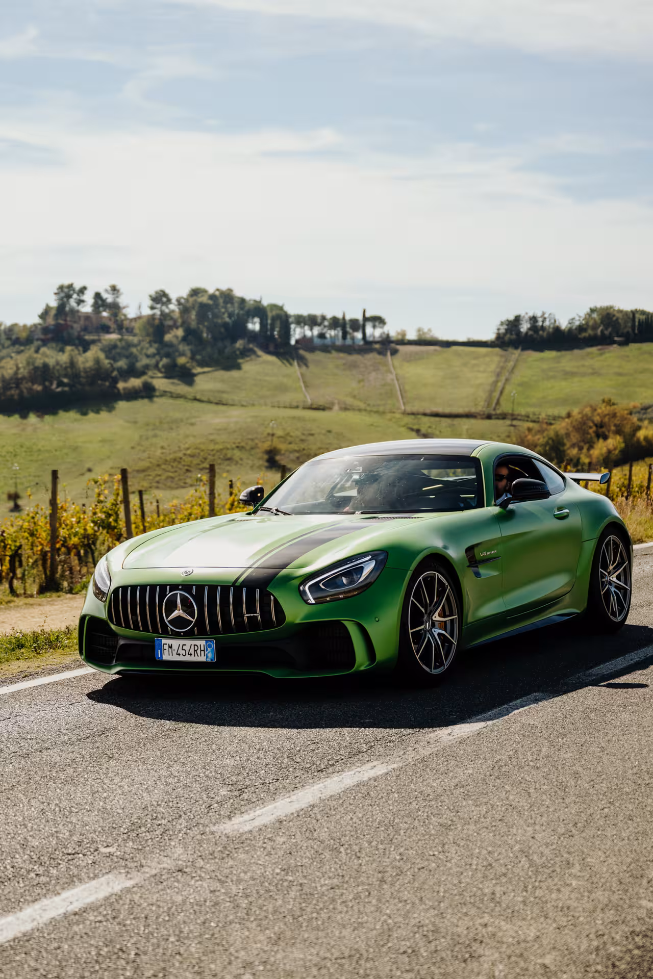 Green Mercedes-AMG GT R sports car on a scenic Tuscan road surrounded by vineyards and rolling hills