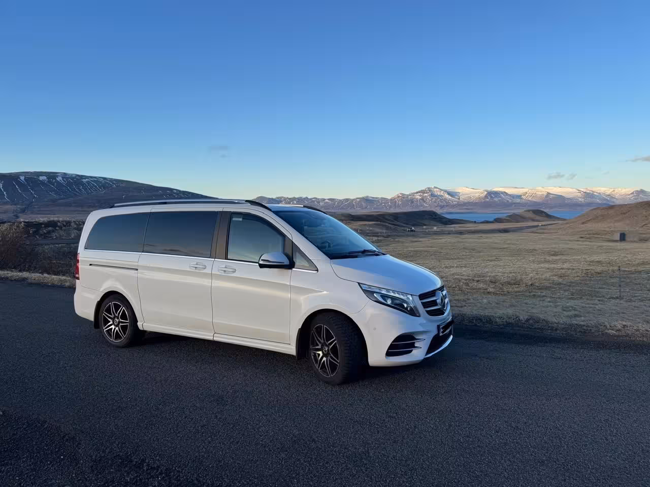 White Mercedes V-Class minivan parked on Icelandic road with snow-capped mountains and fjord in background