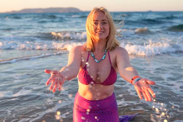 Smiling woman in a purple mermaid tail enjoys a sunset beach photoshoot in Hersonissos, Greece.