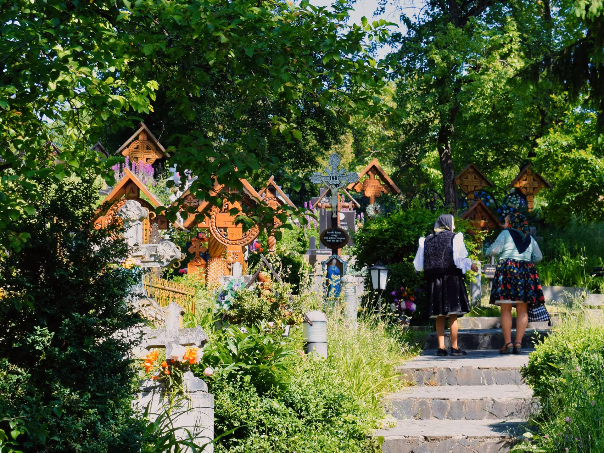 Two women in traditional Romanian folk costumes walk among ornately carved wooden crosses at the Merry Cemetery in Maramures guided tour from Cluj
