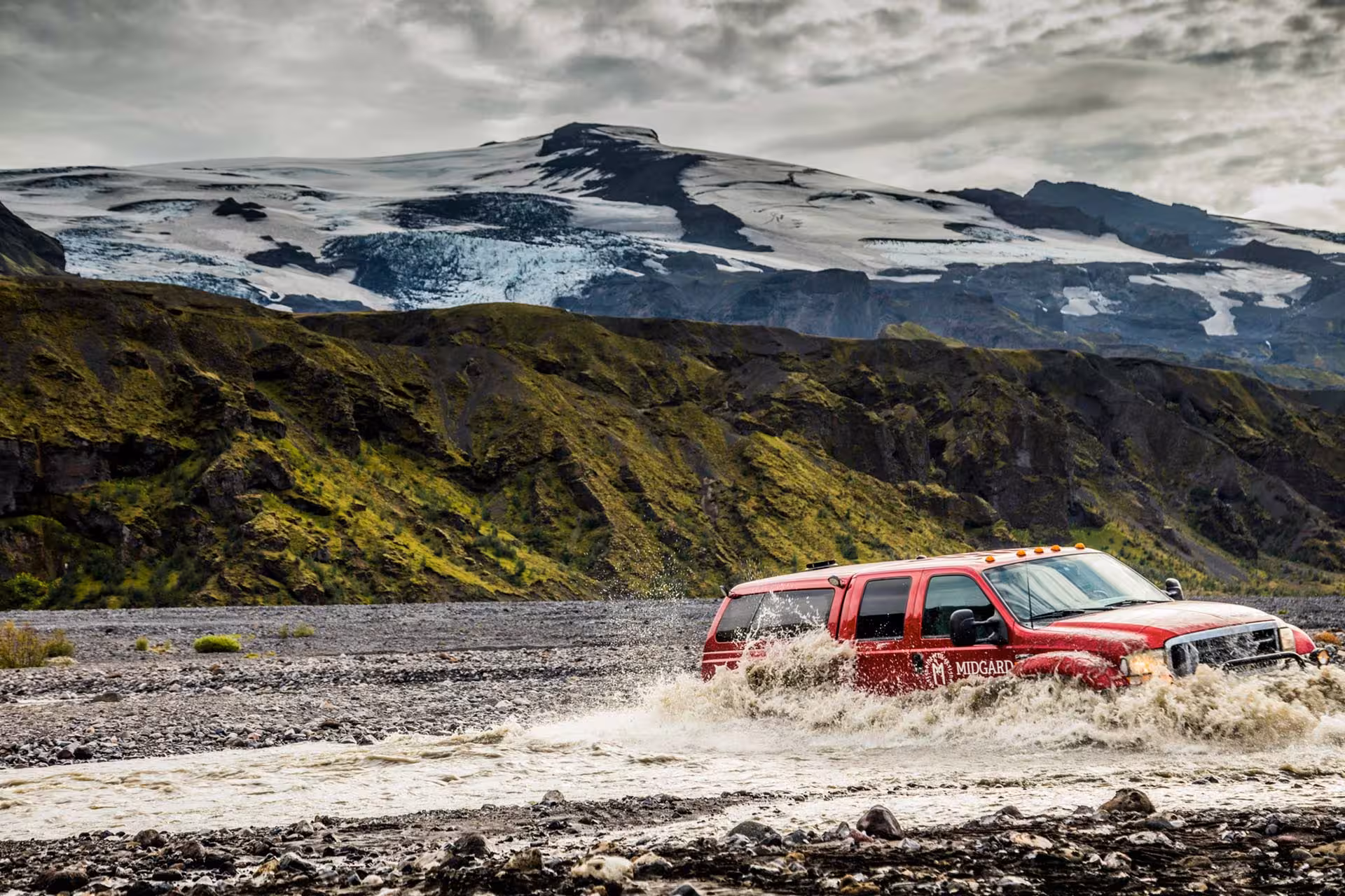 Red Midgard 4x4 SUV crossing glacial river with snow-capped Icelandic mountains in background