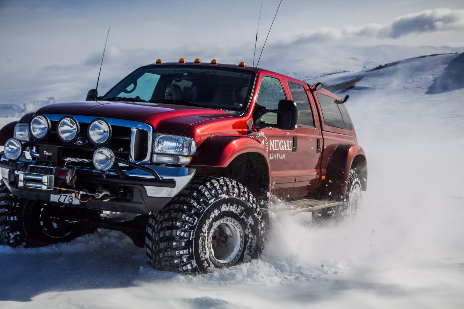 Red Midgard Adventure monster truck driving through snow in Icelandic winter landscape