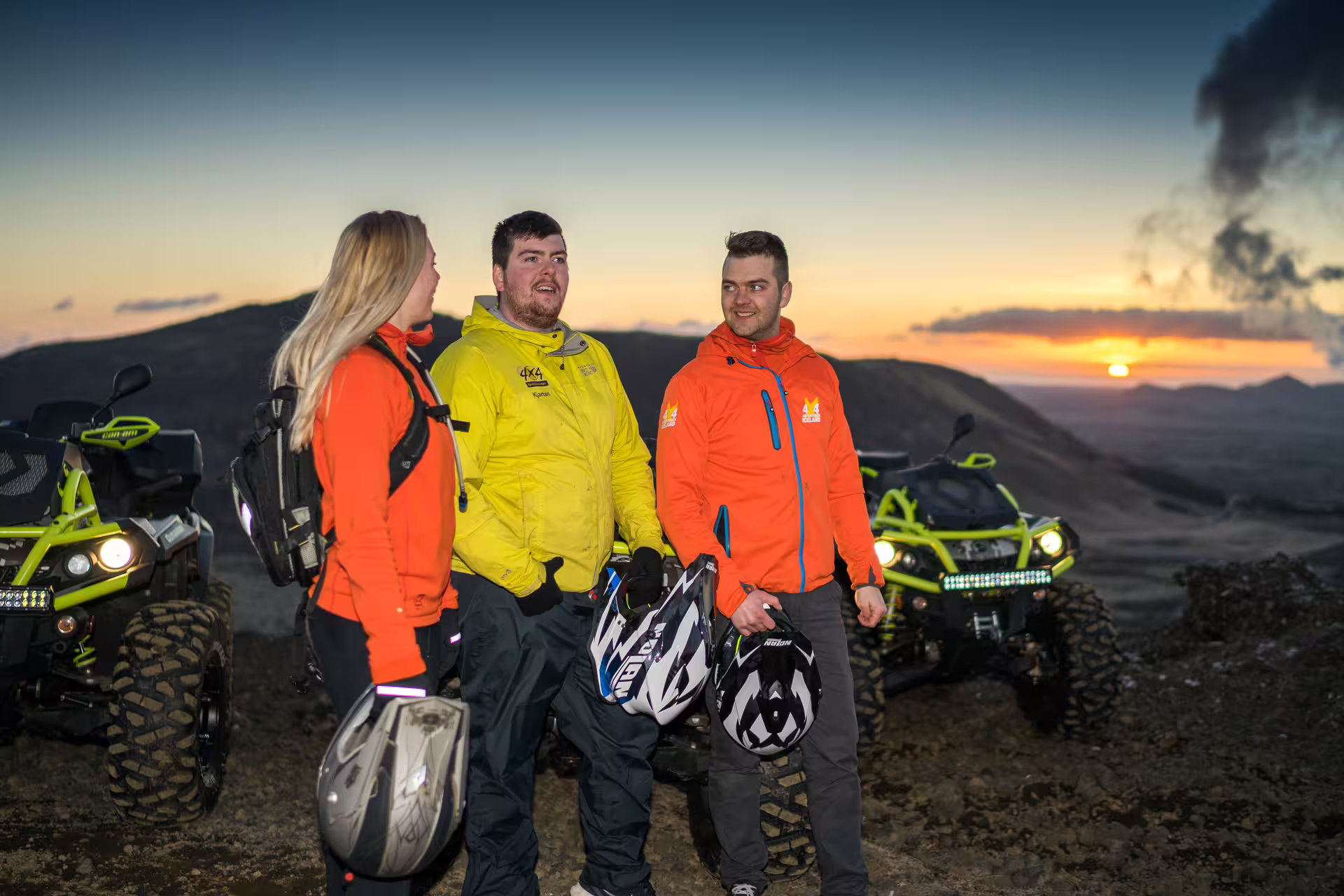 Three people in orange and yellow jackets holding helmets beside ATV quad bikes at sunset in Iceland lava fields