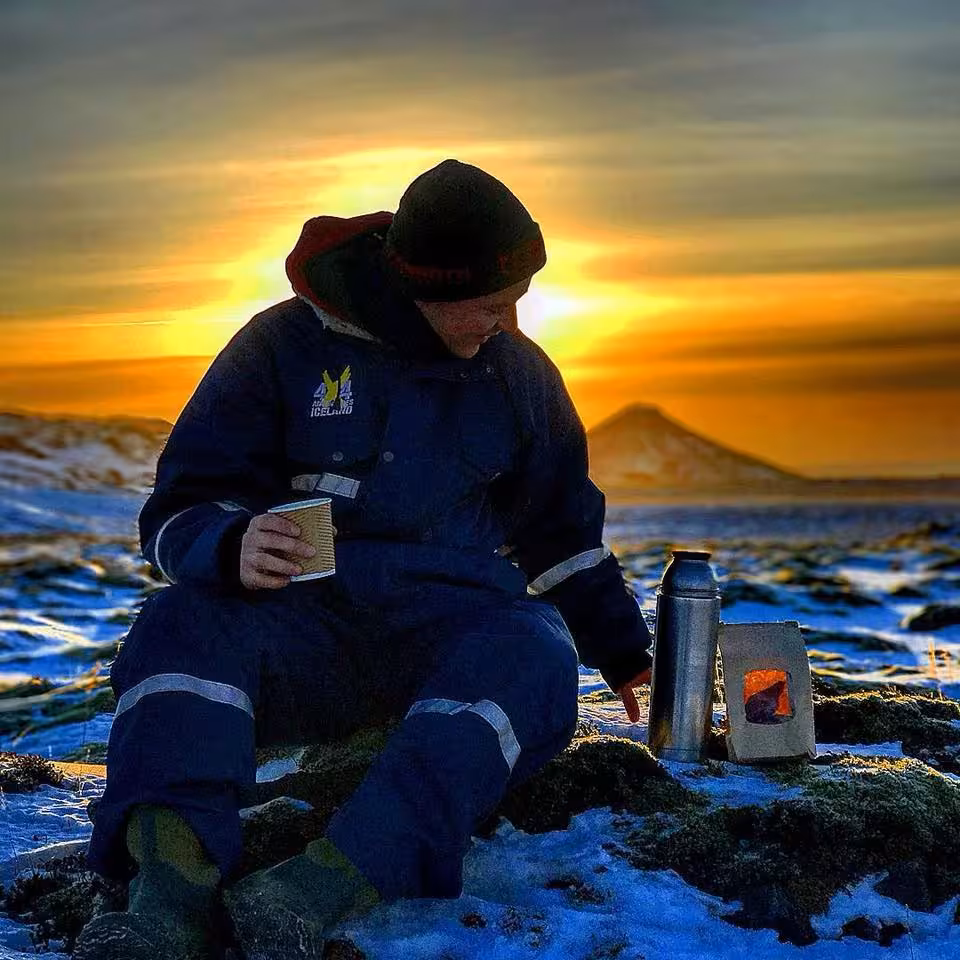 Person in blue overalls drinking hot drink by portable stove during midnight sun in snowy Iceland lava fields