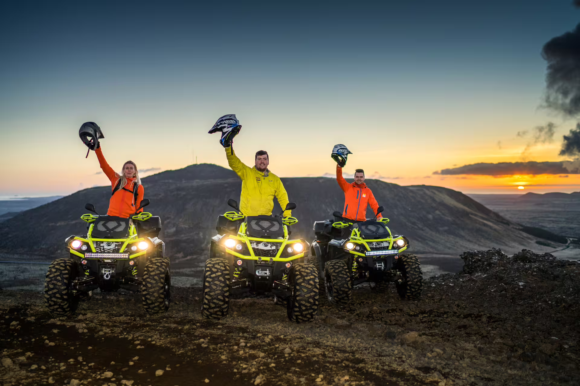 Three riders on neon ATVs raising helmets at sunset over Iceland lava fields volcanic landscape