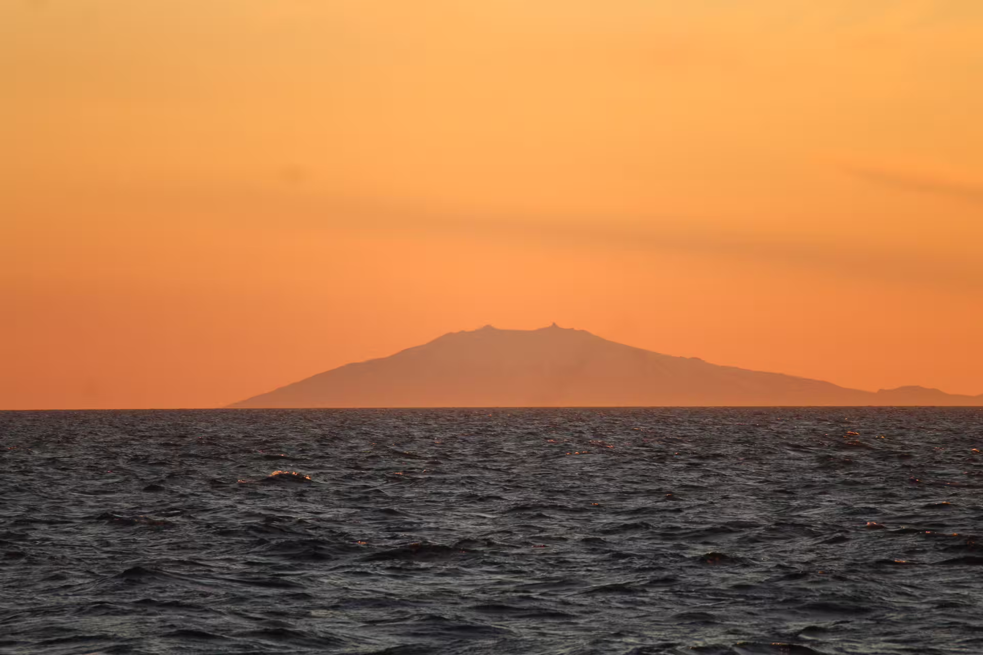 Volcanic island silhouette against vibrant orange midnight sun sky viewed from whale watching boat near Reykjavik