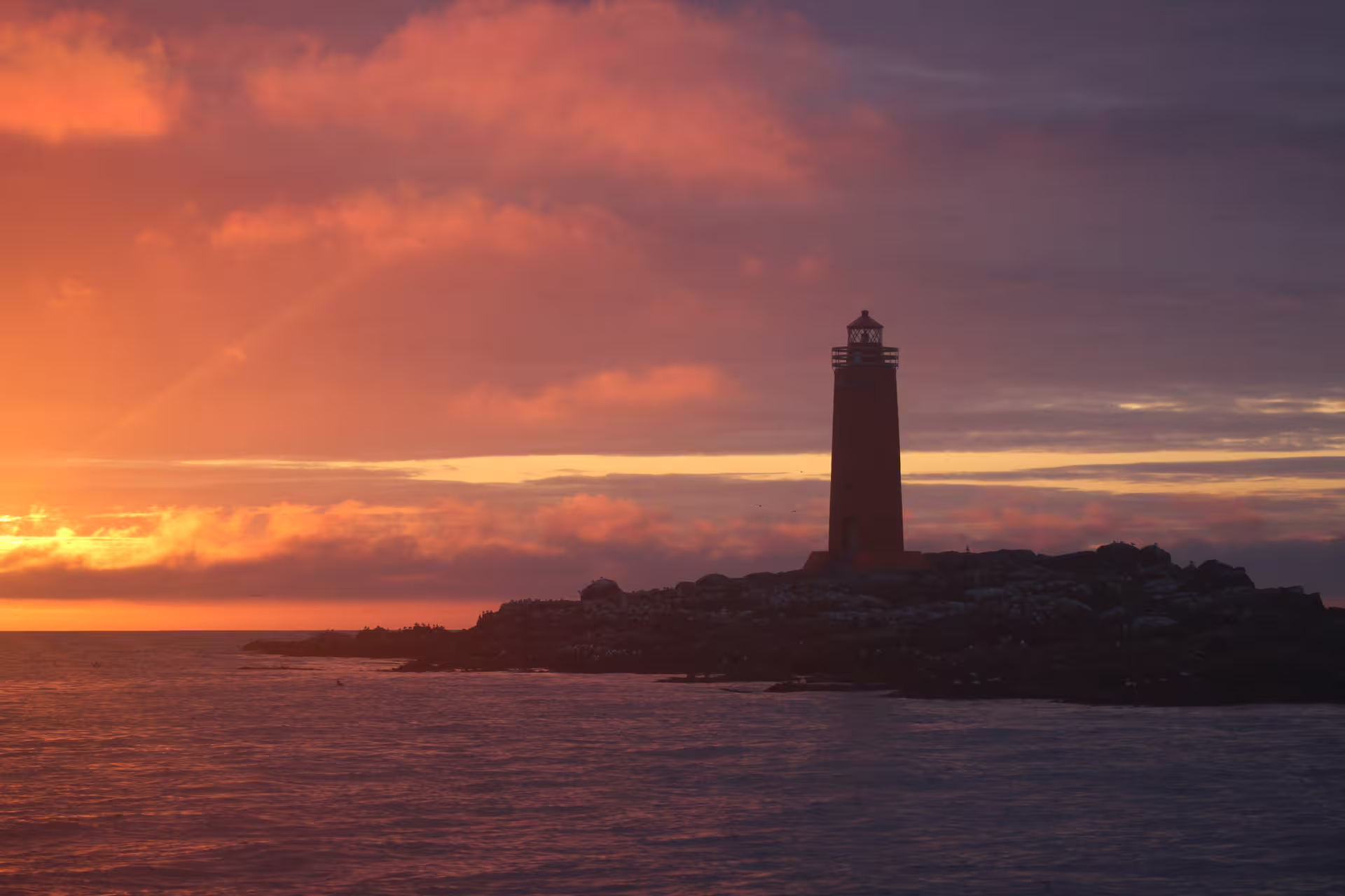 Lighthouse silhouette on rocky island at vivid orange and purple midnight sun sunset over calm ocean waters