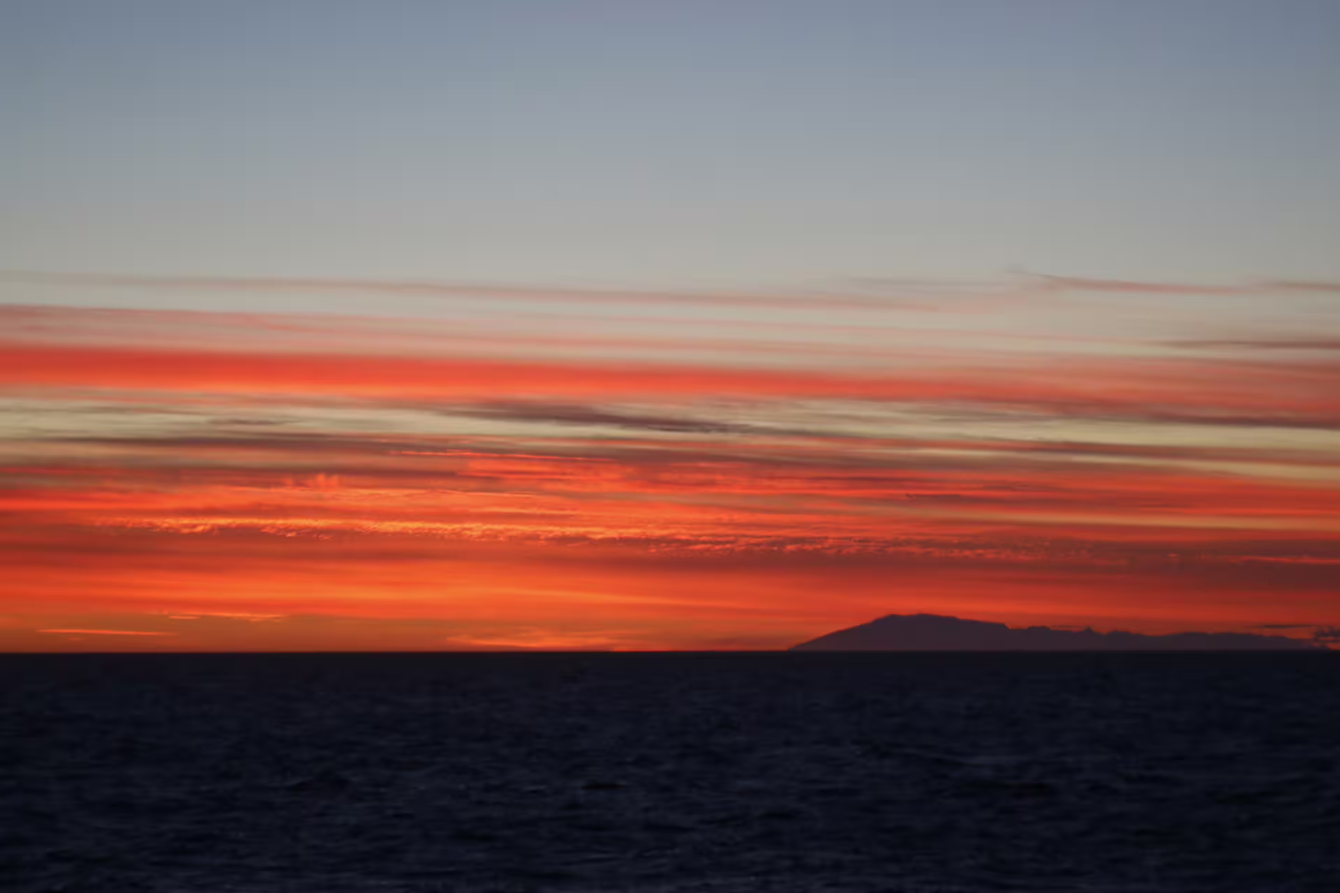 Vivid red and orange midnight sun sky over dark ocean with mountain silhouette on horizon
