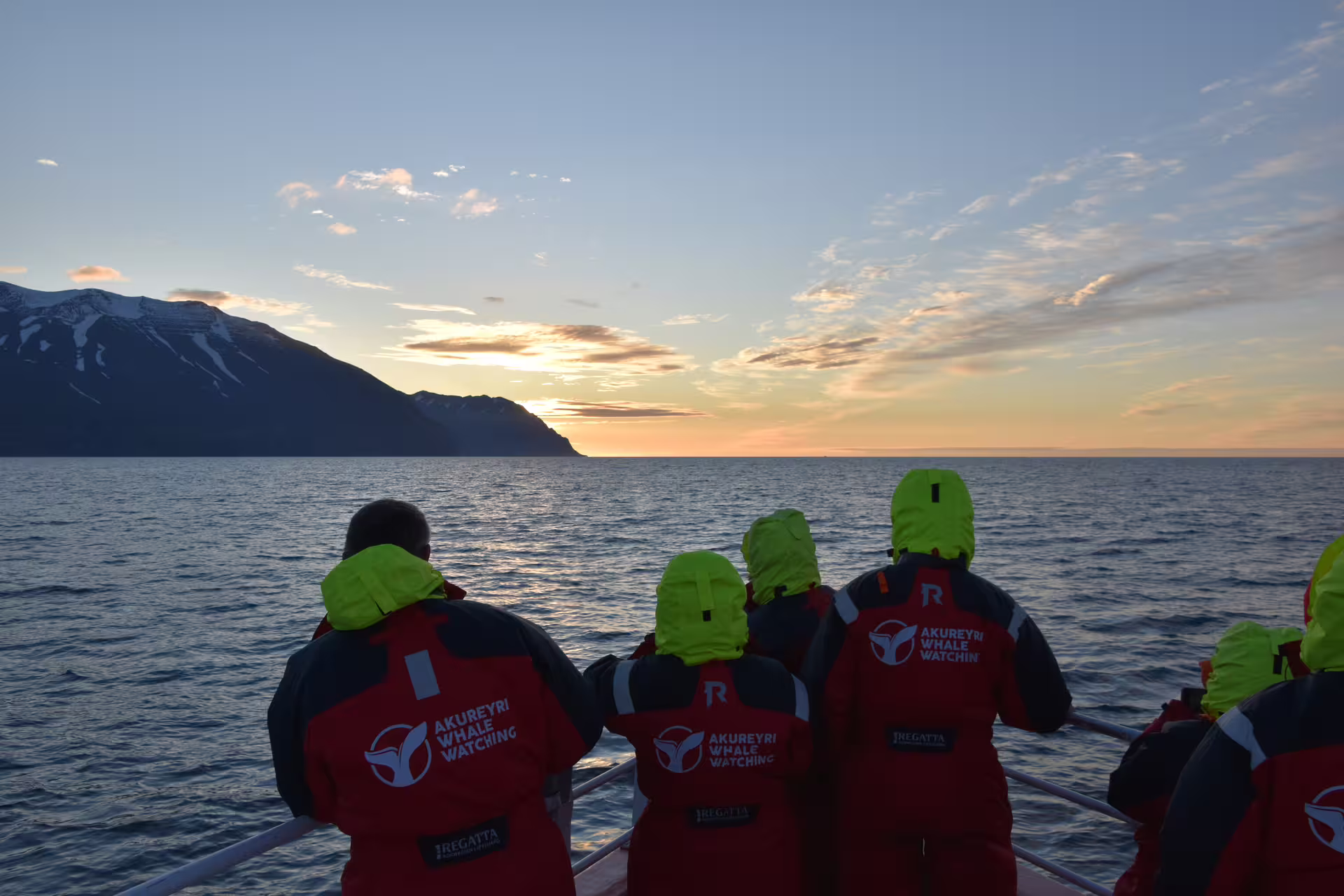 Tourists in red Akureyri Whale Watching jackets watching midnight sun from boat bow in Iceland