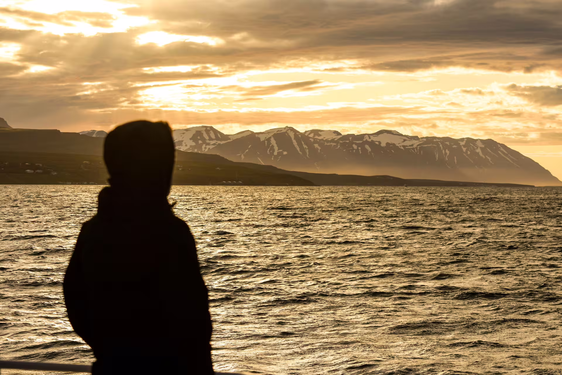 Silhouette of person watching midnight sun over snowy mountains and golden sea in Akureyri