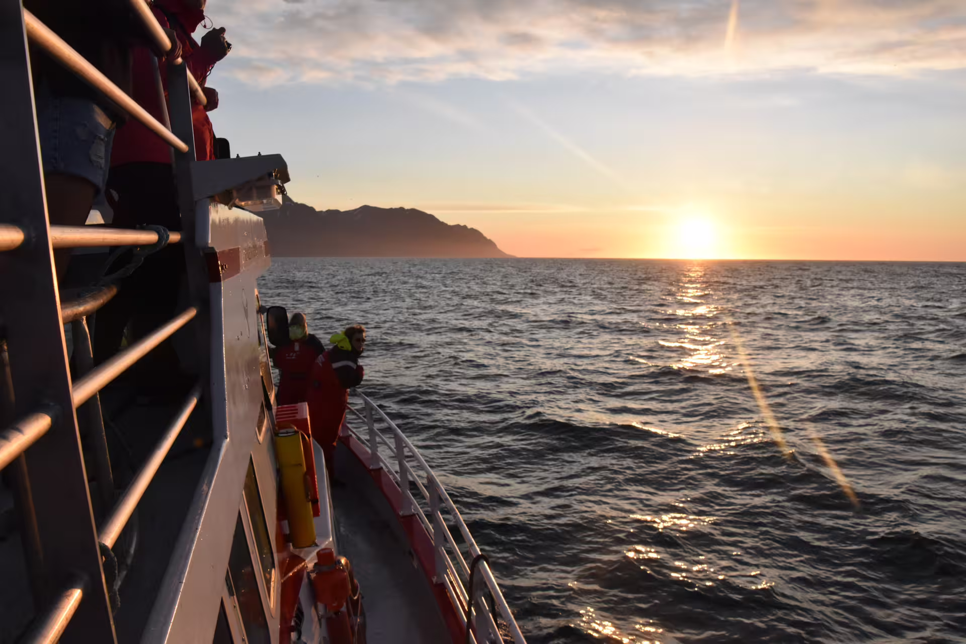 Passengers in red jackets watching midnight sun over calm ocean from whale watching boat near Akureyri