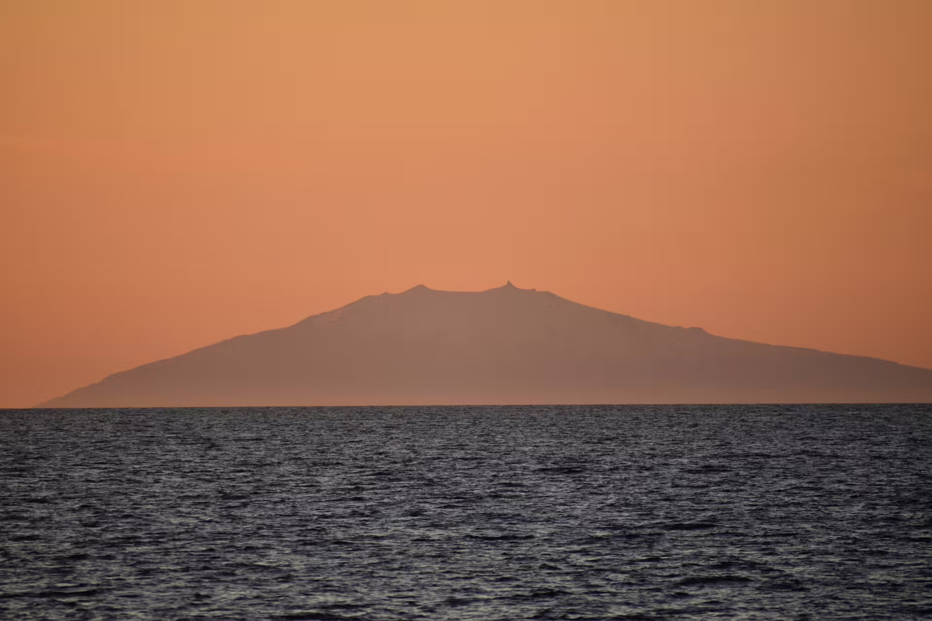 Silhouette of volcanic island over calm ocean under orange midnight sun sky in Reykjavik