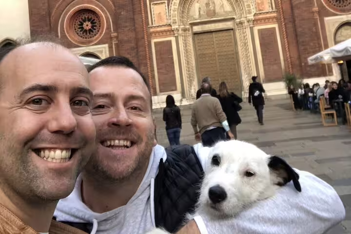 Two smiling men taking a selfie with a white dog in front of a historic Milan church in Brera district
