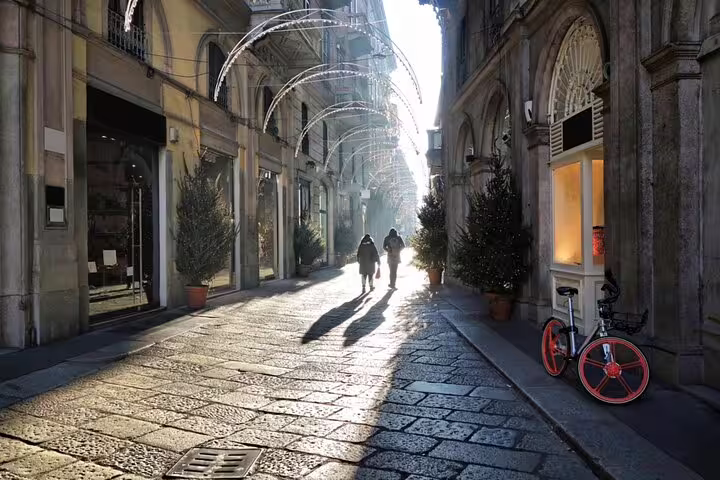 Sunlit historic Milan street with cobblestones, arched lights, and two pedestrians walking between elegant buildings