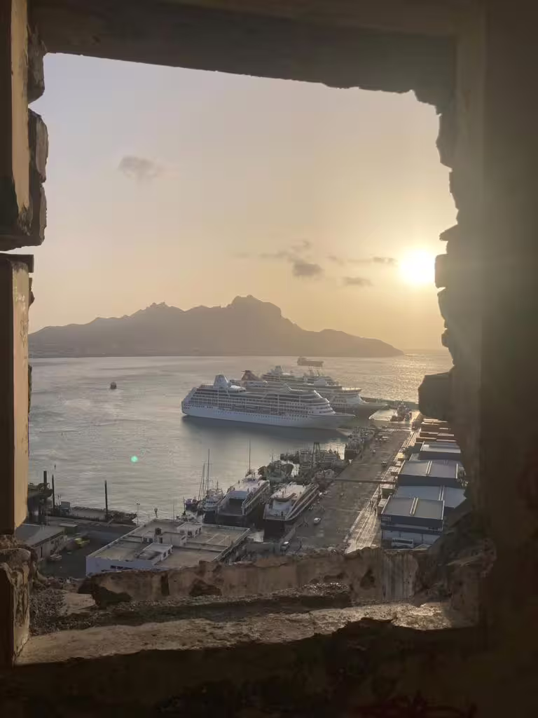 View of a cruise ship docked at Mindelo port during sunset, framed by a historic stone window in São Vicente, Cape Verde.