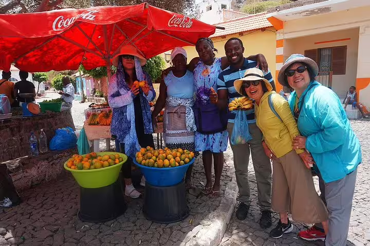 Tourists enjoy fresh local produce at a vibrant fruit market in Mindelo, São Vicente, under a Coca-Cola umbrella.