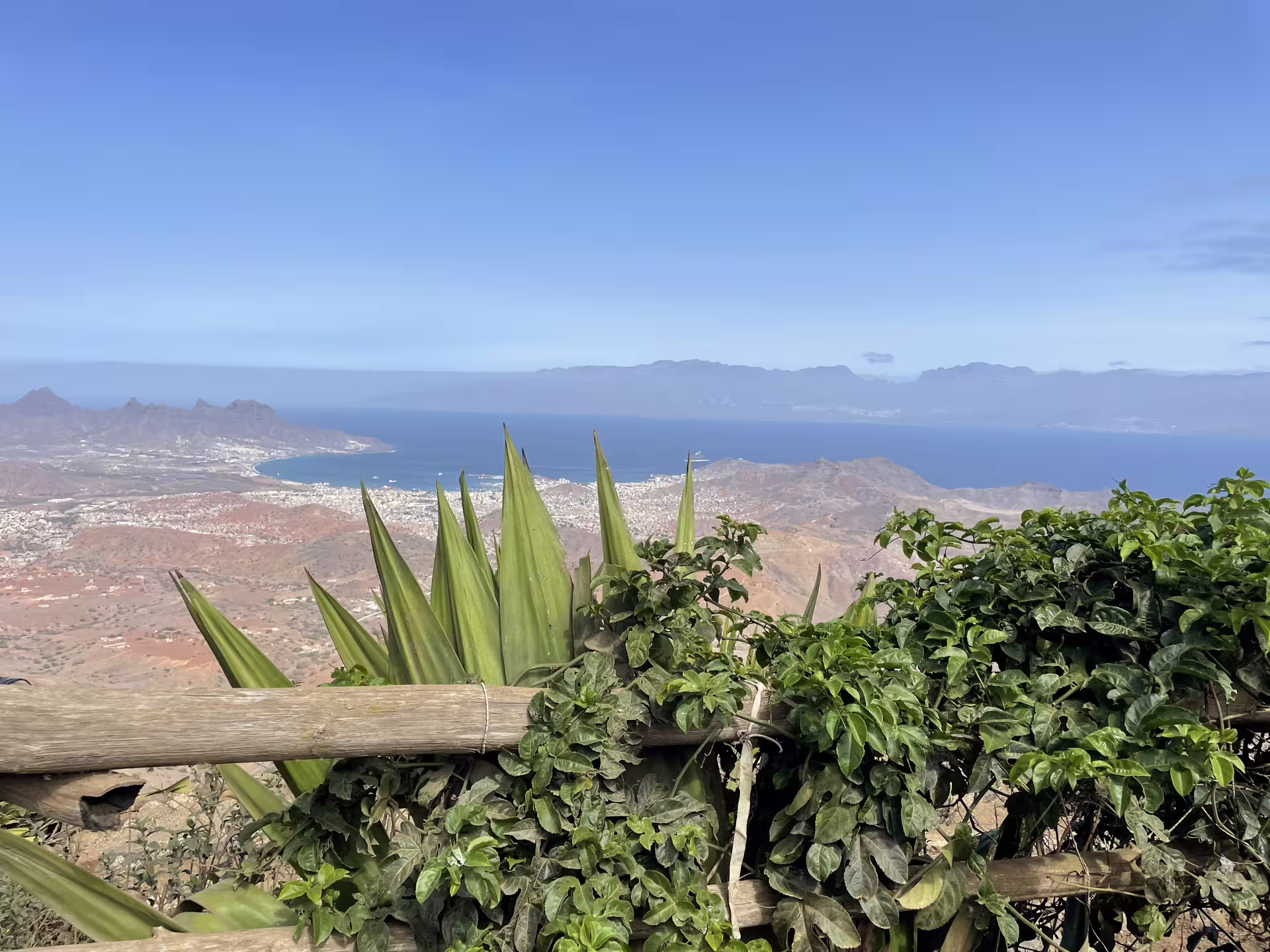 Scenic view of Mindelo's coastline and mountains from São Vicente, showcasing lush greenery and vibrant ocean under clear skies.