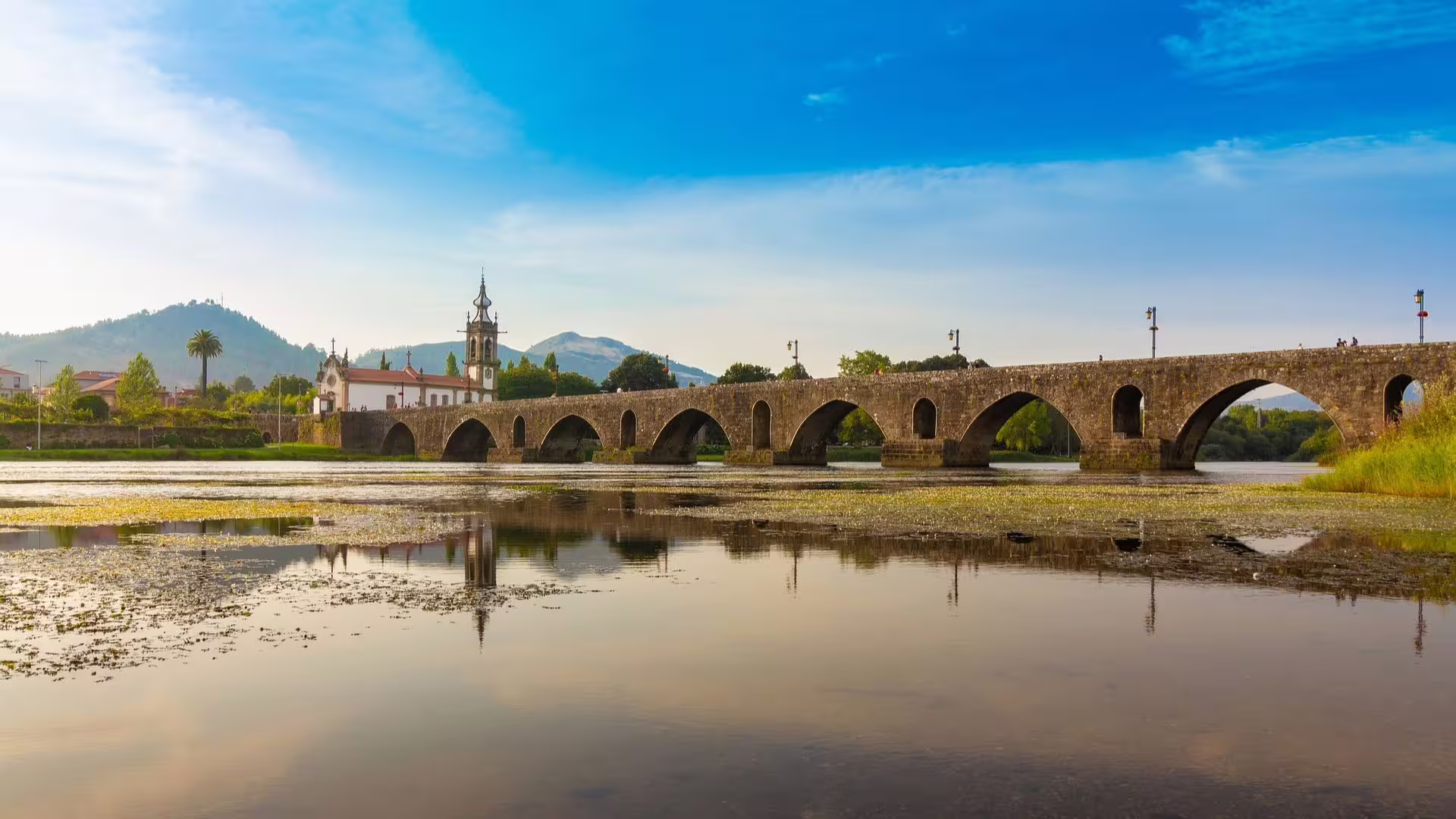 Historic stone bridge over the Lima River in Minho Region, Portugal, reflecting in calm waters under a blue sky.
