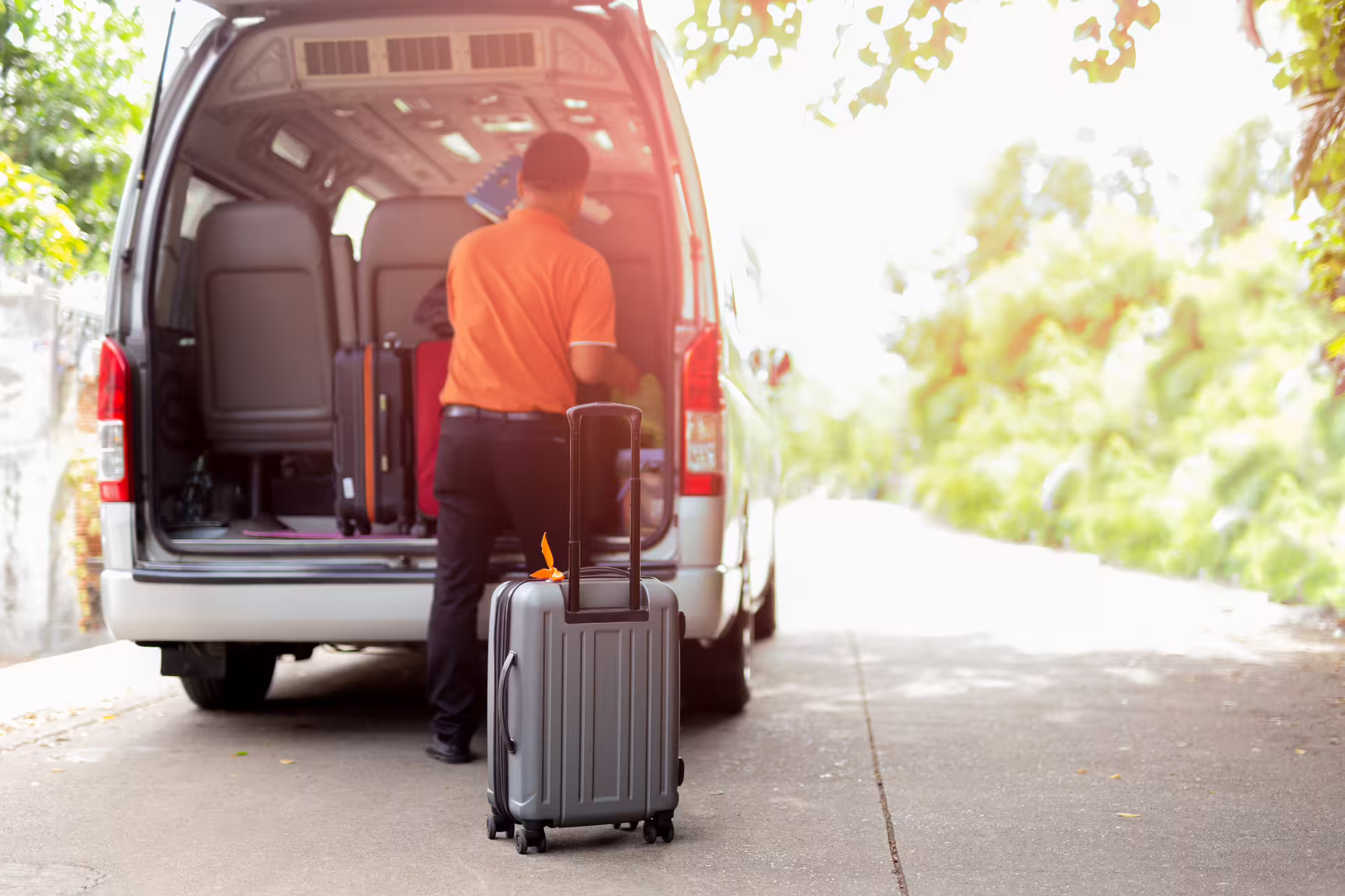Driver in orange shirt loading luggage into minivan transfer vehicle with gray suitcase in foreground