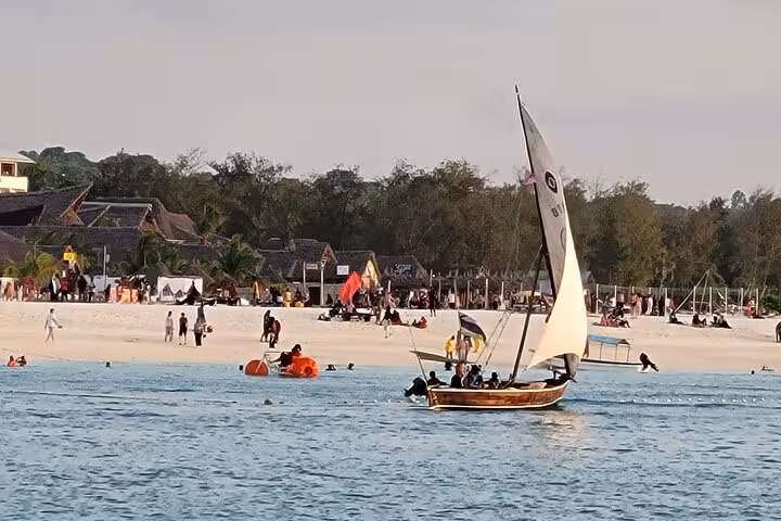 Traditional dhow sailing near a lively Zanzibar beach, part of the Mnemba Island Snorkeling Adventure.