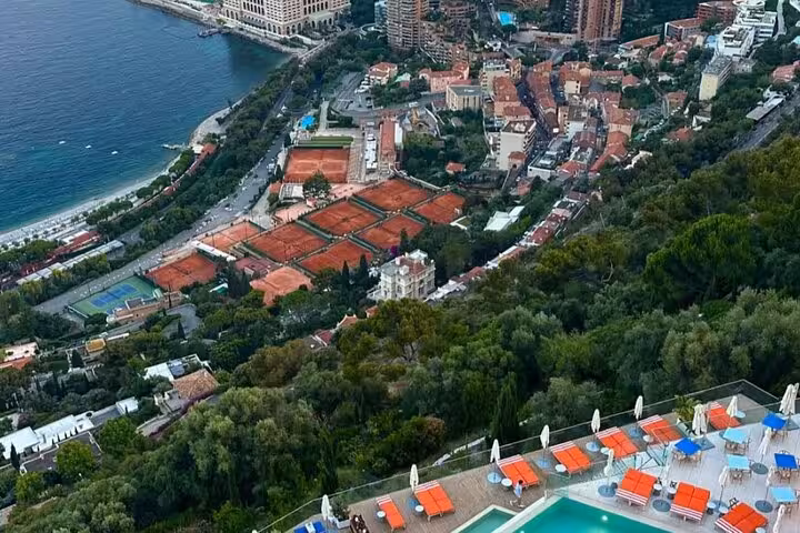 Aerial view of Monaco coastline with red clay tennis courts, dense greenery, and Mediterranean sea