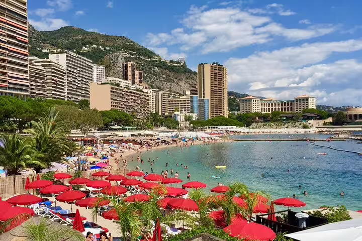 Crowded Monaco beach with red umbrellas, turquoise water, and luxury high-rise buildings backed by green hills