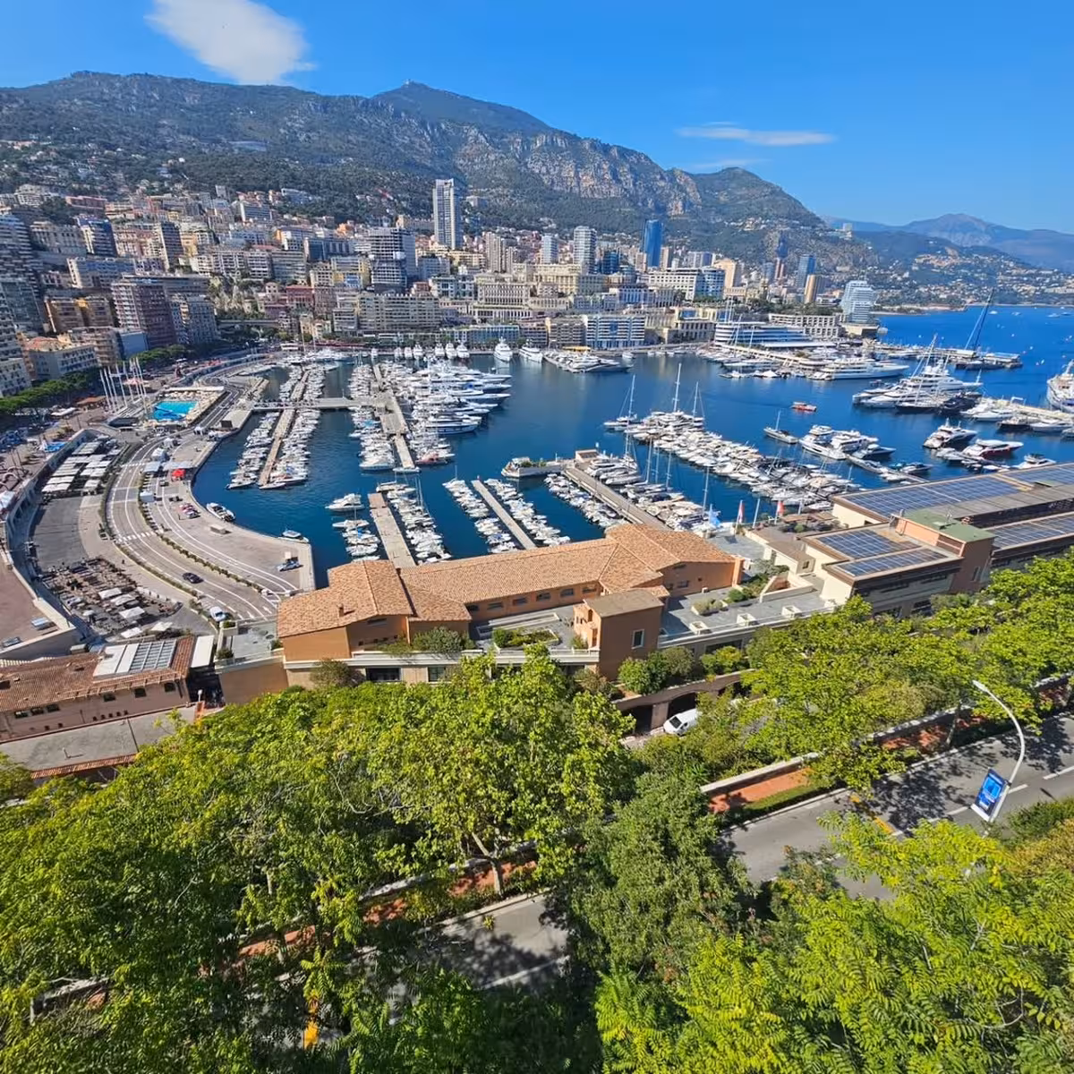 Aerial view of Monaco's Port Hercule marina with luxury yachts, city skyline, and mountain backdrop
