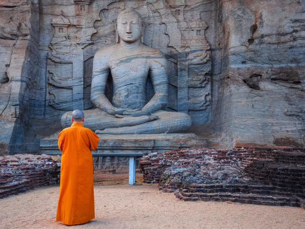 Monk in orange robes prays before ancient Buddha sculpture at Gal Vihara, Polonnaruwa, ideal for Sacred Sri Lanka journeys.