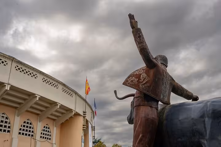 Bronze bullfighter statue raising hand outside Mont de Marsan bullring with Spanish flag under cloudy sky