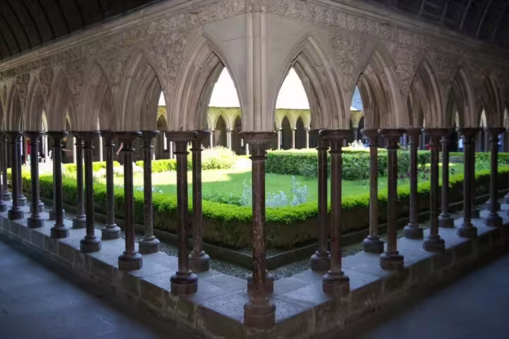 Gothic cloisters with ornate arched columns surrounding a lush green garden courtyard at Mont Saint-Michel Abbey