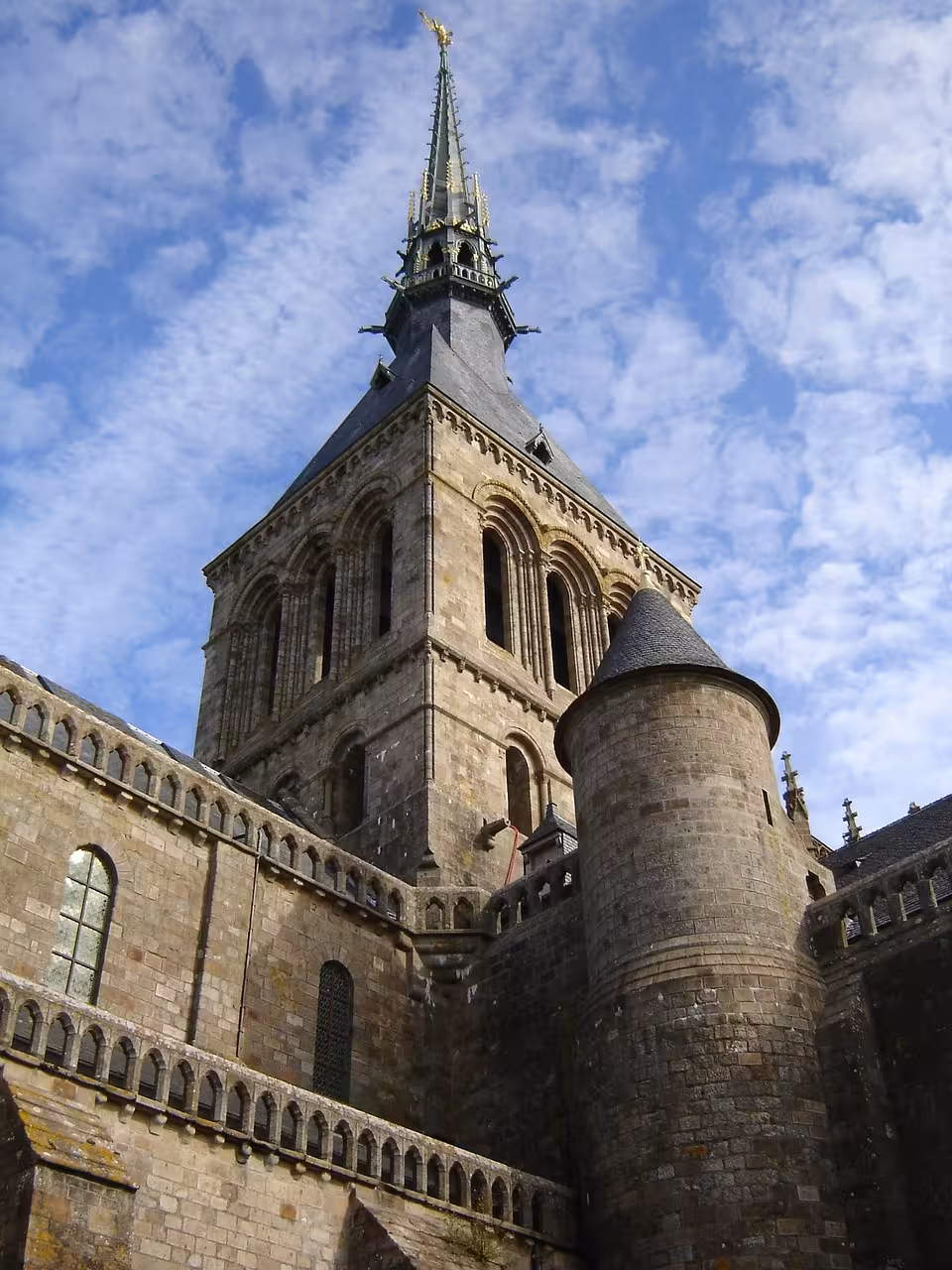 Gothic spire and stone tower of Mont Saint-Michel abbey seen from below against blue sky