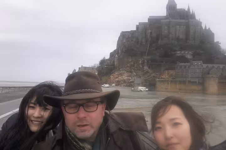 Three tourists taking a selfie with Mont-Saint-Michel abbey rising on rocky island behind them
