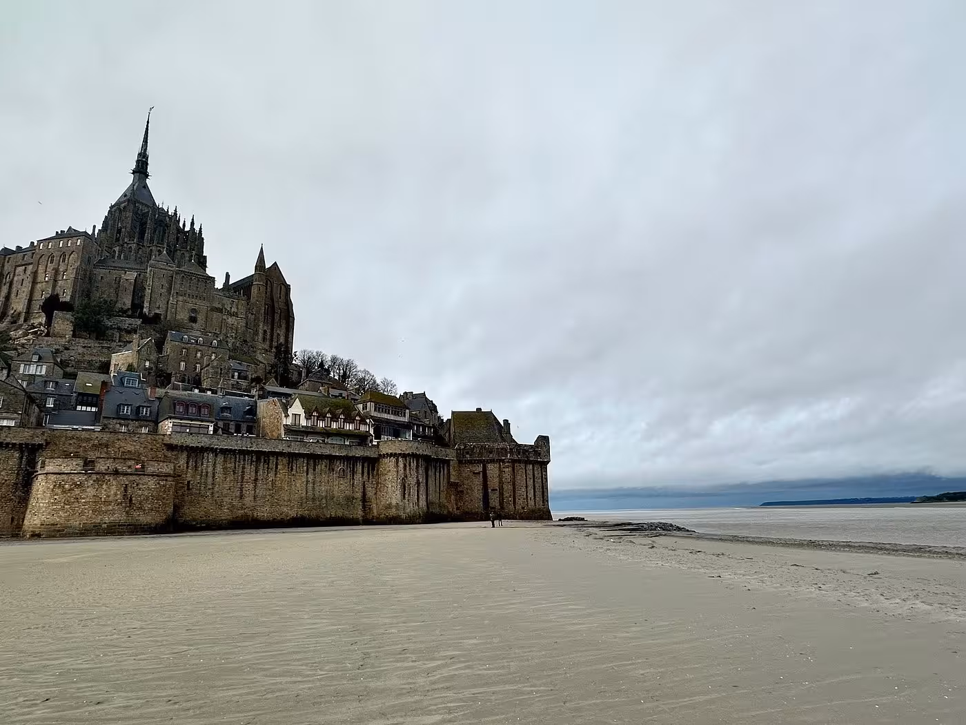 Mont-Saint-Michel abbey and medieval walls viewed from tidal sand flats at low tide under overcast sky
