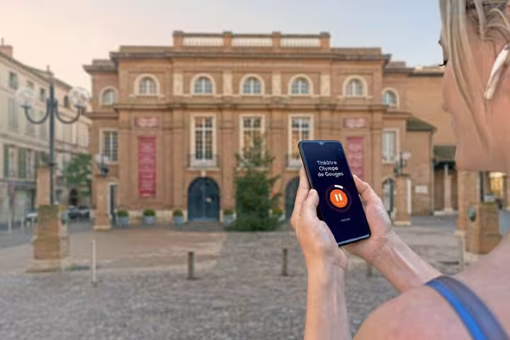 Woman holding smartphone with audio tour app in front of Théâtre Olympe de Gouges in Montauban